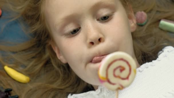 Klein meisje met een lolly ligt op een blauwe achtergrond. Close-up portret, bovenaanzicht — Stockvideo