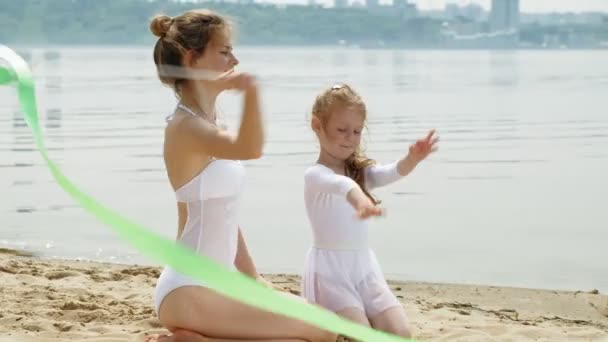 Madre e hija en trajes de baño blancos bailando con cinta de gimnasia en una playa de arena. Verano, amanecer — Vídeo de stock