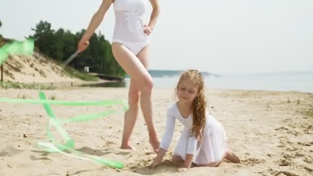 Mãe e filha em trajes de banho brancos dançando com fita ginástica em uma praia de areia. Verão, amanhecer — Vídeo de Stock