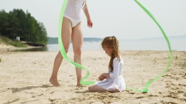 Ibu dan anak dalam pakaian renang putih menari dengan pita senam di pantai berpasir. Musim panas, fajar — Stok Video