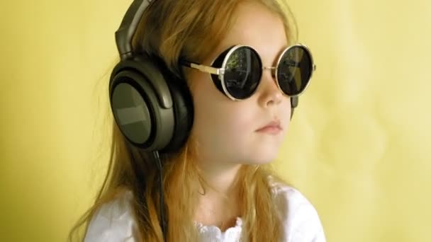 黄色の背景にヘッドフォンで陽気な小さな女の子。クローズアップポートレート. — ストック動画