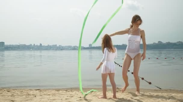 Mãe e filha em trajes de banho brancos dançando com fita ginástica em uma praia de areia. Verão, amanhecer — Vídeo de Stock