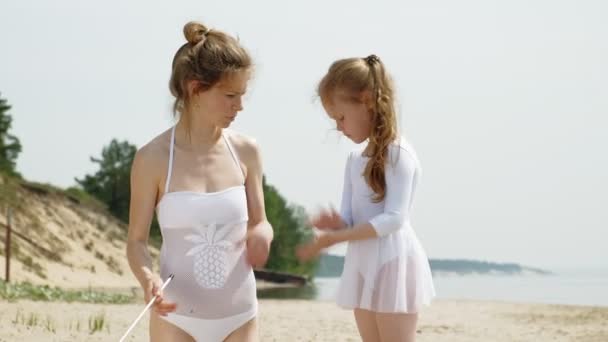 Mother and daughter in white bathing suits dancing with gymnastic ribbon on a sandy beach. Summer, dawn — Stock Video