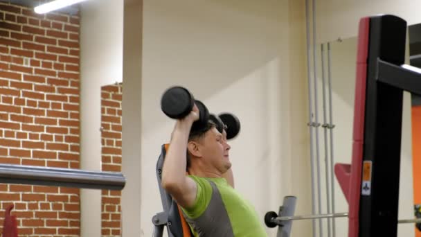 Un hombre haciendo un press de banca con pesas sentado con un agarre recto en un gimnasio — Vídeo de stock