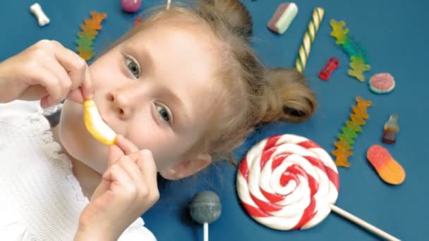 Ein fröhliches kleines Mädchen liegt mit Süßigkeiten auf blauem Grund. Nahaufnahme-Porträt — Stockvideo