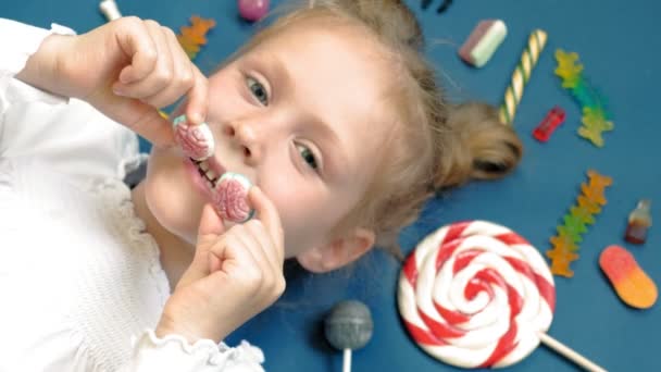 Niña alegre se encuentra sobre un fondo azul con dulces. Retrato de primer plano — Vídeos de Stock