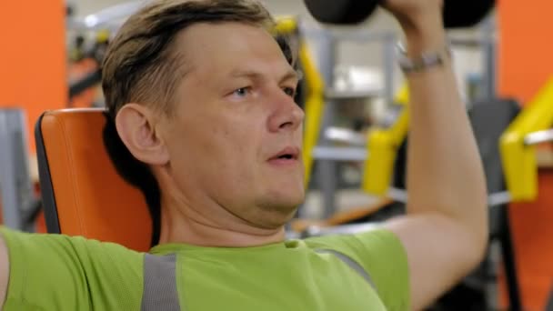 En man gör en bänkpress med hantlar sitter med ett rakt grepp i en fitness Studio — Stockvideo