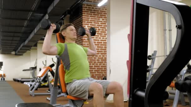 Человек, делающий пресс для скамеек с гантелями, сидящий с прямой хваткой в фитнес-студии — стоковое видео
