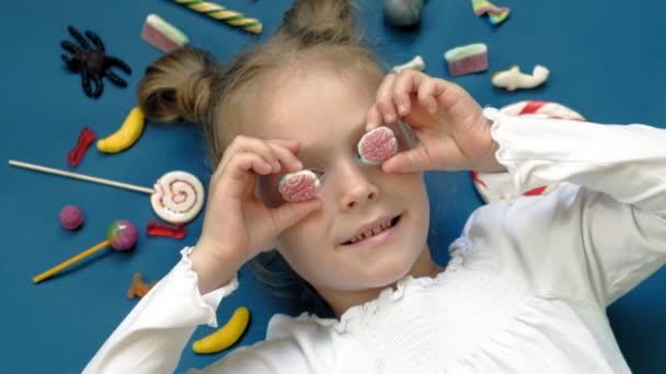 Χαρούμενο κοριτσάκι βρίσκεται σε ένα μπλε φόντο με γλυκά. Κοντινό πλάνο — Αρχείο Βίντεο
