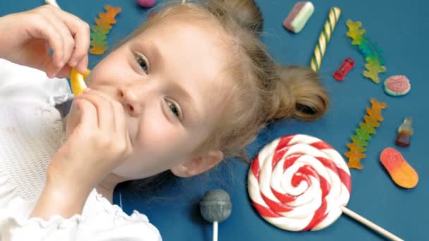 Niña alegre se encuentra sobre un fondo azul con dulces. Retrato de primer plano — Vídeos de Stock