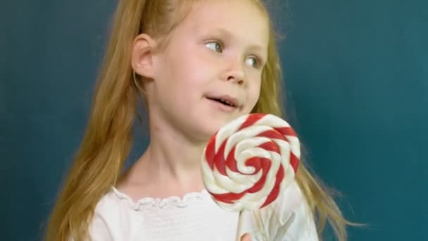 Niña con una piruleta sobre un fondo azul. Primer plano retrato — Vídeos de Stock