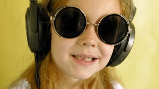 Fröhliches kleines Mädchen mit Kopfhörern auf gelbem Hintergrund. Nahaufnahme-Porträt. — Stockvideo