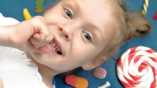 Ein fröhliches kleines Mädchen liegt mit Süßigkeiten auf blauem Grund. Nahaufnahme-Porträt — Stockvideo