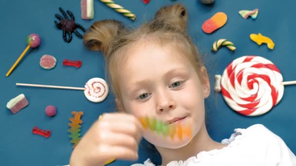Весела маленька дівчинка лежить на синьому фоні з цукерками. Портрет крупним планом — стокове відео