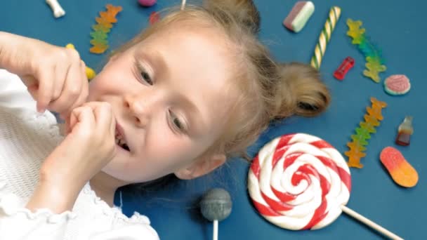 Neşeli küçük kız tatlılar ile mavi bir arka plan üzerinde yatıyor. Yakın çekim portre — Stok video