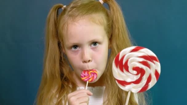 Klein meisje met een lolly op een blauwe achtergrond. Close-up staand — Stockvideo