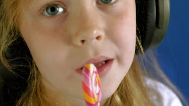 Ένα κοριτσάκι με γλειφιτζούρι σε μπλε φόντο. Κοντινό πορτρέτο — Αρχείο Βίντεο