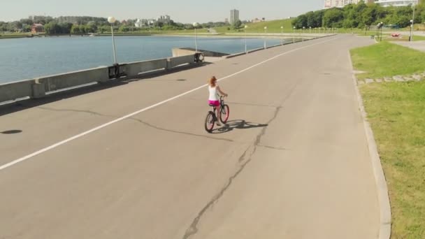 Mujer joven montando una bicicleta al aire libre en verano. Terraplén del río. Transporte ecológico. Disparo aéreo — Vídeos de Stock