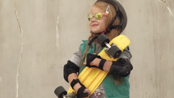 Una niña alegre se para con un patín amarillo en las manos y sonríe — Vídeos de Stock