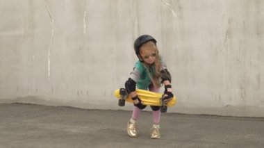 Neşeli bir küçük kız elinde sarı bir paten ile duruyor ve gülümsüyor
