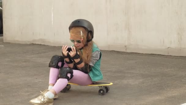 Niña alegre está sentada en un monopatín amarillo y está usando un teléfono móvil. Navegación por Internet — Vídeo de stock