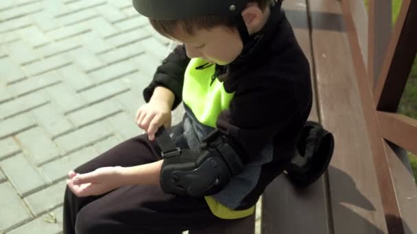 Un enfant d'âge préscolaire s'assoit sur un banc dans le parc et se protège la tête, les genoux et les bras. Été — Video