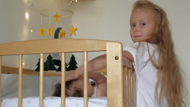 Дівчинка дошкільного віку стоїть на ліжку з дитиною, заспокоює її. Догляд за маленькою сестрою — стокове відео