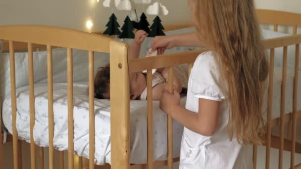 Vorschulmädchen steht mit Kind an der Krippe, beruhigt sie. Pflege einer kleinen Schwester — Stockvideo