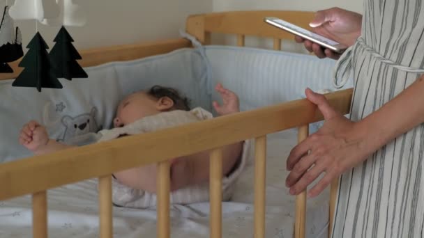 The kid plays with the mobile in the crib. Mom stands by the bed and uses the phone. — Stock Video