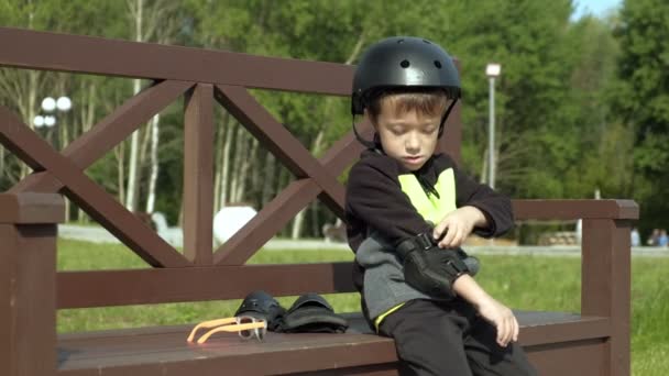 Un niño en edad preescolar se sienta en un banco del parque y se protege la cabeza, las rodillas y los brazos. Verano — Vídeos de Stock