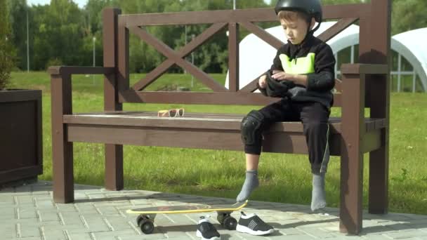 Ένα νήπιο κάθεται σε ένα παγκάκι στο πάρκο και βάζει προστασία στο κεφάλι, τα γόνατα και τα χέρια του. Καλοκαίρι — Αρχείο Βίντεο