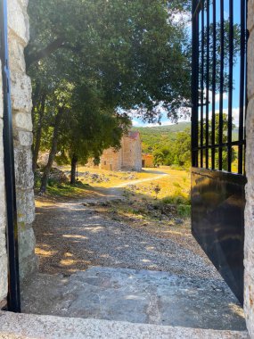Yunanistan 'da ağaçlar ve tepelerle çevrili eski taştan bir kiliseye açılan açık demir bir kapıdan bak. Bir yaz gününde tarihi mimari ve sıcak güneş ışığıyla huzurlu bir kır manzarası..