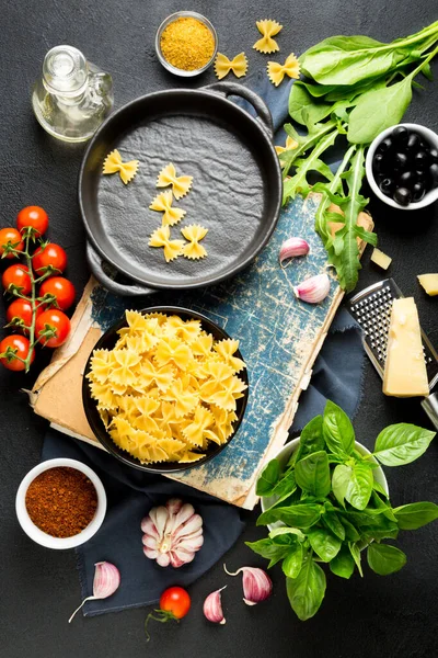 Makarna malzemeleri. Kiraz domatesleri, farfalle makarna, sarımsak, fesleğen, parmesan, ıspanak, roka, baharat ve zeytinyağı ve koyu bir zemin. Üst görünüm