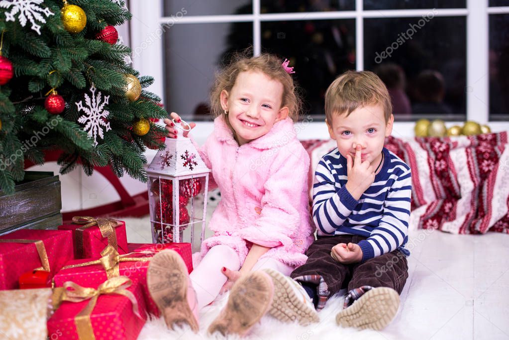 Two small children, brother and sister, are sitting on the background of the Christmas interior and gifts. Merry Christmas and Happy Holidays! Christmas tree indoors.