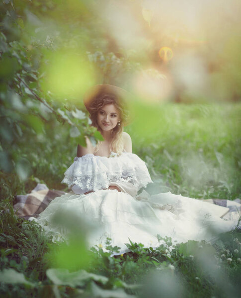 Beautiful girl in a white vintage dress and a hat sits on the grass