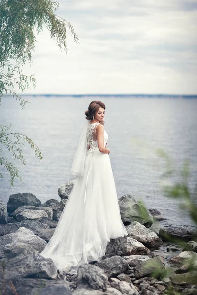 La novia en un vestido blanco en las piedras cerca del río — Foto de Stock