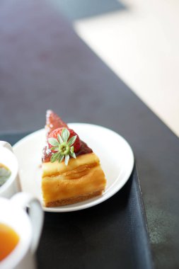 Dilim peynir Kek krem ve taze çilek plaka üzerinde bulanıklık arka plan üzerinde bir fincan çay ile kek. Çay saati kavramı Seçici odak