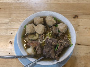 Rahatlatıcı bir kase Endonezya bakso 'sunun tadını çıkarın. İçinde lezzetli et suyu, sulu biftek kaburgaları, sert köfteler ve ahşap bir masada sarı erişteler var.