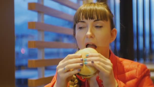 Mujer comiendo comida rápida. Retrato de una joven caucásica mordiendo y masticando hamburguesa con queso en un restaurante de comida rápida . — Vídeos de Stock