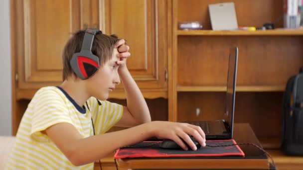 Tiener met behulp van de laptop. Tiener jongen met koptelefoon vastgelijmd aan het laptopscherm met behulp van de muis van het gokken van de computer en hoofd krabben. — Stockvideo