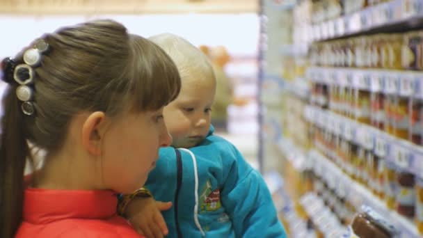 Atractiva mujer caucásica eligiendo comida infantil en el supermercado sosteniendo al bebé en brazos. Primer plano de mamá y bebé mirando purés de frutas en los estantes de la tienda de comestibles. Niño recoge un frasco de comida para bebés . — Vídeos de Stock