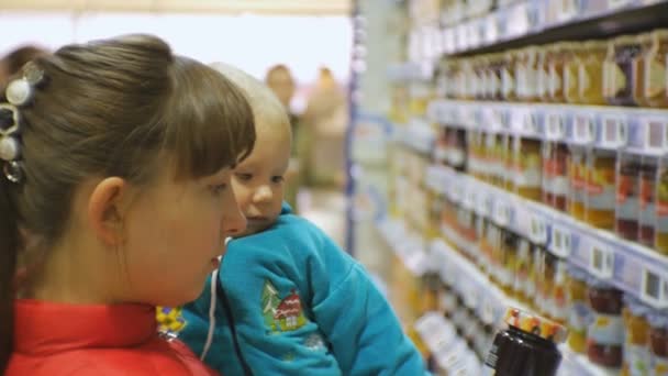 Attraente donna caucasica che sceglie il cibo per bambini al supermercato tenendo in braccio il bambino. Un primo piano di mamma che guarda il vasetto del cibo per bambini in mano alle madri. Donna che legge l'etichetta . — Video Stock