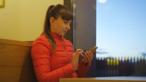 Una mujer atractiva en el café. Retrato de mujer caucásica en chaqueta de burbuja roja tocando la pantalla de su teléfono inteligente cerca de una ventana en la noche . — Vídeo de stock