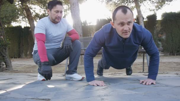 Προπόνηση με προσωπικό γυμναστή σε εξωτερικούς χώρους. Γυμναστήριο άνθρωπος κάνει σούπερ αργή push-ups σε ένα πάρκο ως μέρος της μια ρουτίνα προπόνηση. Ο προπονητής παρακολουθεί μετρώντας τα δευτερόλεπτα — Αρχείο Βίντεο