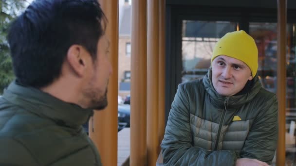 Primer plano de dos atletas masculinos en chaquetas de burbuja caliente charlando después de entrenar en el área al aire libre de la cafetería por la noche . — Vídeos de Stock