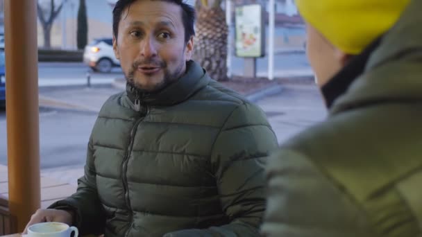 Dos atletas masculinos en cálidas chaquetas de burbuja charlando después de entrenar en el área al aire libre de la cafetería por la noche. Primer plano del tipo con la barba dice algo sobre el entrenamiento de hoy . — Vídeos de Stock