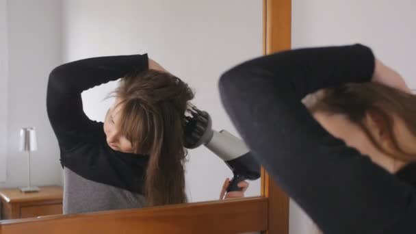 Giovane attraente donna dai capelli castani gira la testa asciugando i capelli con un asciugacapelli davanti allo specchio . — Video Stock