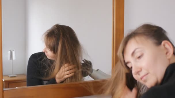 Jeune jolie femme aux cheveux bruns sourit séchant ses cheveux avec un sèche-cheveux devant le miroir à la maison . — Video