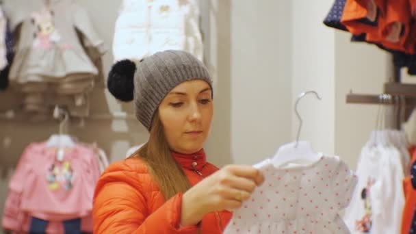 Attraktiv kvinna i baby kläder butik. Kaukasiska kvinna i grått sticka mössa och röd jacka håller baby klänning i händer prövningen. — Stockvideo