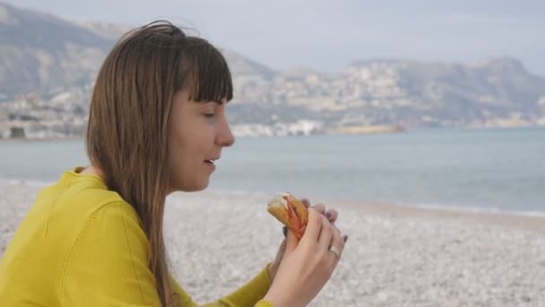 Retrato de una joven hembra comiendo en la playa. Atractiva mujer caucásica mordiendo sándwich en la playa fuera de temporada . — Vídeo de stock